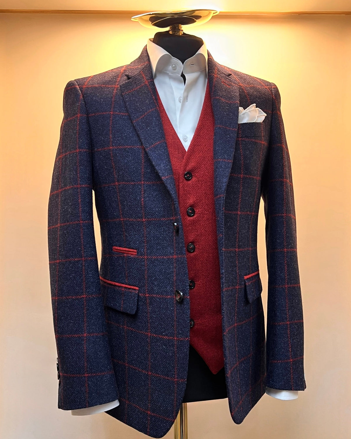 A navy and red check blazer on a mannequin, with a red waistcoat in the background, both against a beige wall.