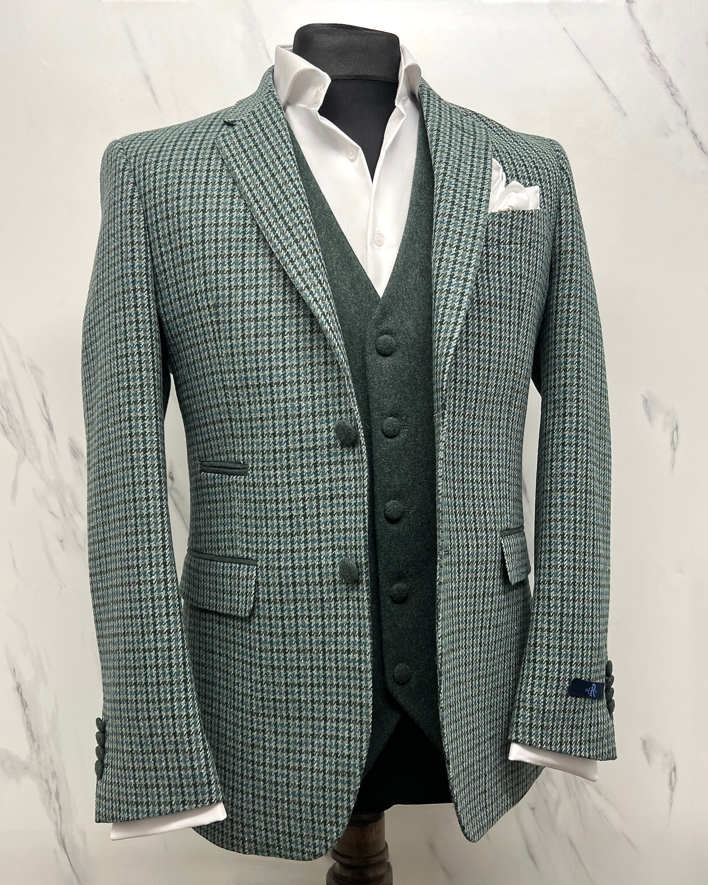 A sage green houndstooth blazer with a notch lapel, paired with a white shirt and a green waistcoat, displayed on a mannequin against a marble background.
